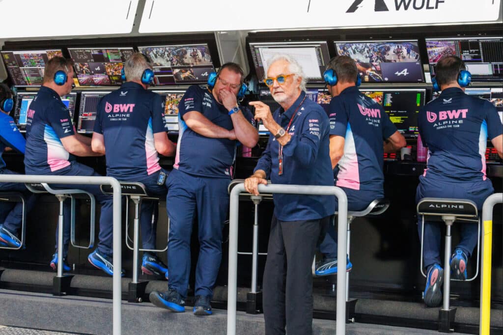 A medium shot of Flavio Briatore, Alpine's Executive Advisor and de facto team principal, standing in the Formula One paddock. He is wearing his signature blue-tinted sunglasses, a white dress shirt, and a dark blazer. His expression is serious and direct, reflecting his reputation for decisive authority and high-stakes decision-making within the Enstone-based team.