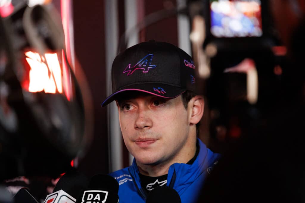 A close-up, high-intensity photograph of Franco Colapinto wearing a black Alpine team cap with pink and blue detailing and a blue team jacket. He is surrounded by microphones from various media outlets, including DAZN, during a press interview. His expression is serious and focused, captured in a dimly lit environment with warm bokeh lights in the background.