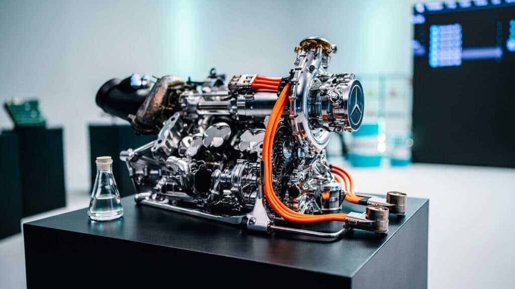 A highly detailed, close-up photograph of a Mercedes-Benz Formula One power unit displayed on a black pedestal in a clean, laboratory-like environment. The engine features complex metallic components, a visible Mercedes star emblem on the side, and prominent orange high-voltage cables. In the background, a glass beaker and digital displays add to the high-tech, engineering-focused atmosphere.