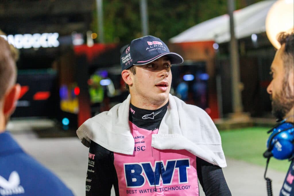 A close-up, profile photograph of Franco Colapinto seated in the cockpit of his Alpine F1 car. He is wearing a white and blue racing helmet with "BWT" and "Alpine" branding, his dark eyes visible and focused through the clear visor. The background is blurred, showing the carbon fiber halo and the top of the blue Alpine chassis, emphasizing the intense concentration required for the 2026 season.