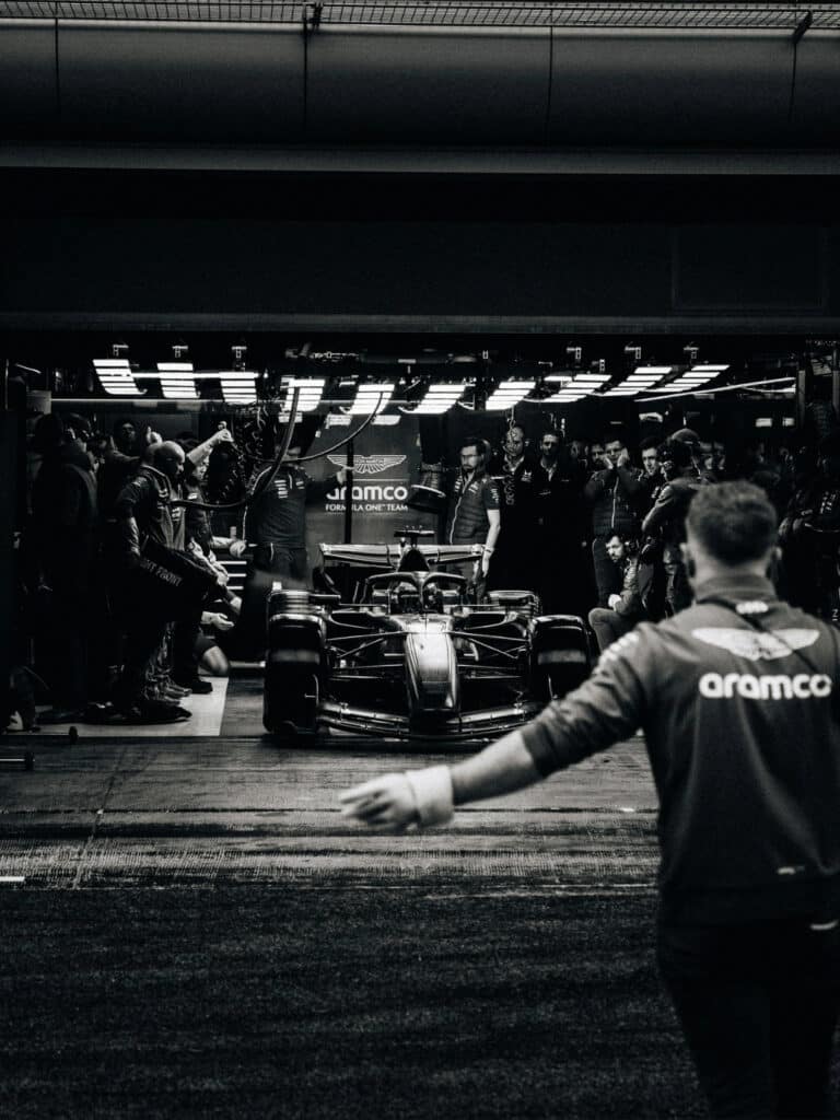 Aston Martin Aramco F1 car under pit garage lights with crew members preparing for a session, capturing the high-pressure atmosphere of the 2026 regulation cycle.