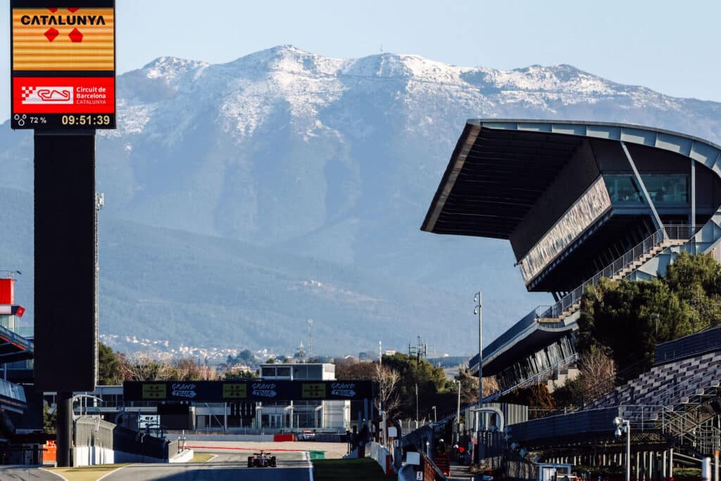 A Formula 1 car on the main straight of the Circuit de Barcelona-Catalunya with snow-capped mountains in the background during 2026 testing.