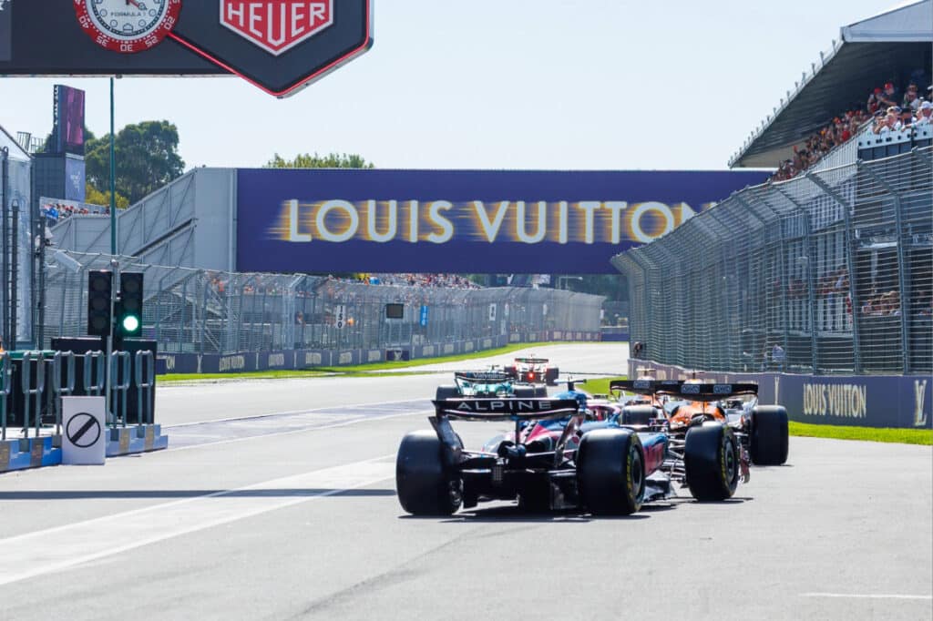 Formula 1 cars racing through the Albert Park Circuit in Melbourne with packed grandstands and the Louis Vuitton bridge in the background.