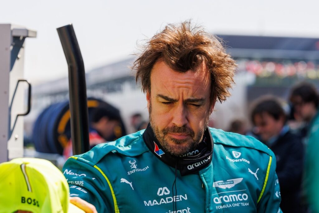A candid shot of Fernando Alonso in his Aston Martin racing suit, looking downcast and weary during a race weekend, highlighting the team's reliability struggles in 2026.