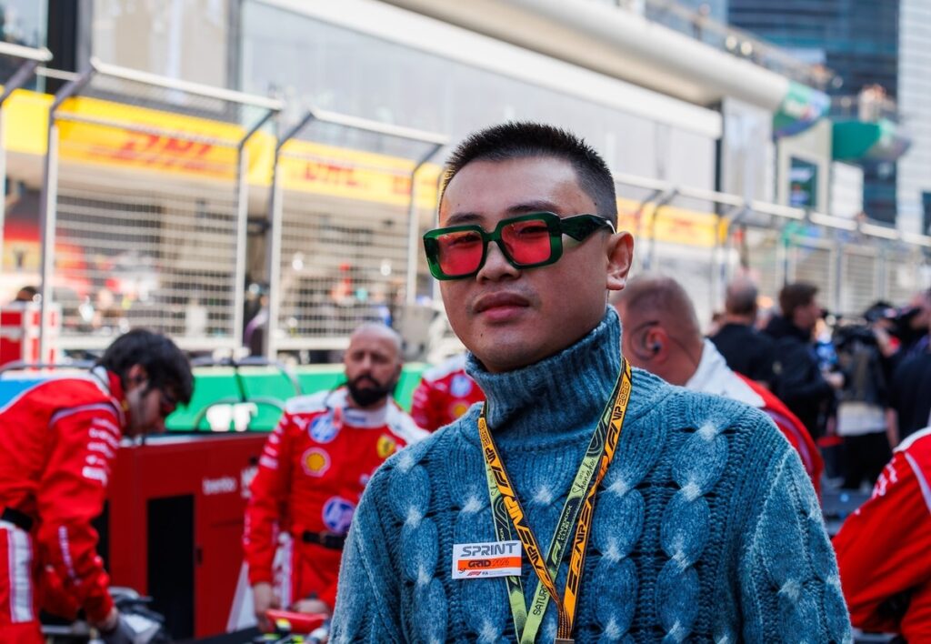 Chinese singer and actor Huo Zun wearing a blue sweater and red-tinted sunglasses in the F1 paddock at the 2026 Chinese Grand Prix.