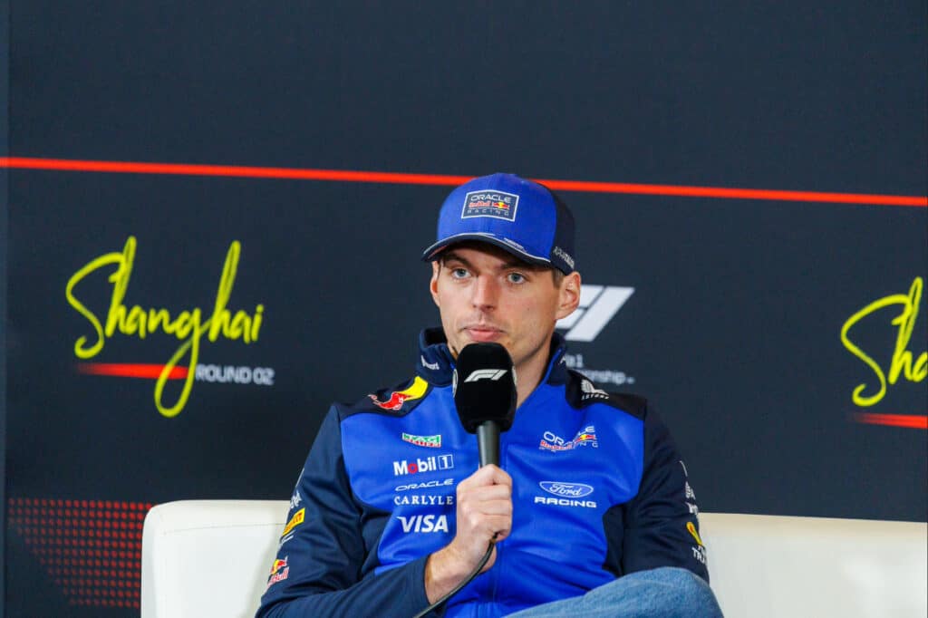 Max Verstappen speaking into a microphone during the FIA press conference at the 2026 Chinese Grand Prix, discussing the new Formula One technical regulations.